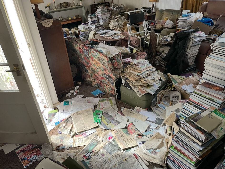 A cluttered living room with stacks of papers, magazines, and books everywhere, including on the floor, furniture, and other surfaces.