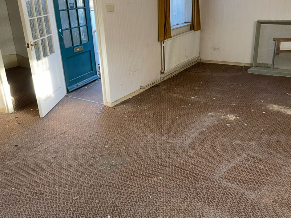 Empty room with stained brown carpet, white walls, blue door, window with yellow curtains, and scattered debris on the floor.