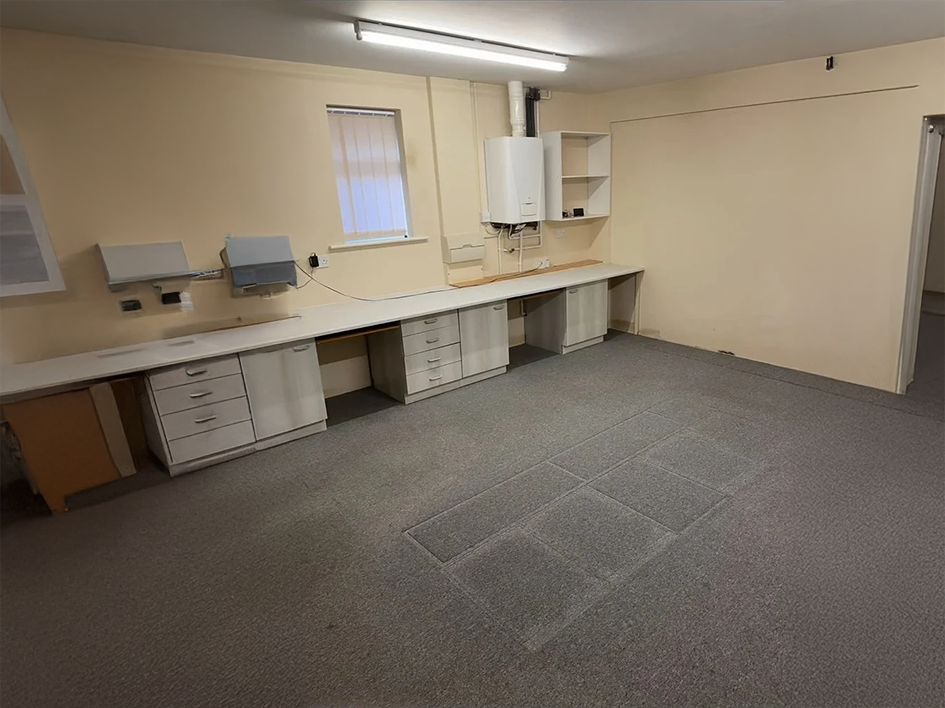 Empty office or laboratory space with beige walls, gray carpet, a long white counter with multiple drawers, two wall-mounted paper towel dispensers, a window with vertical blinds, a white wall-mounted heater or boiler, and shelving on the wall.