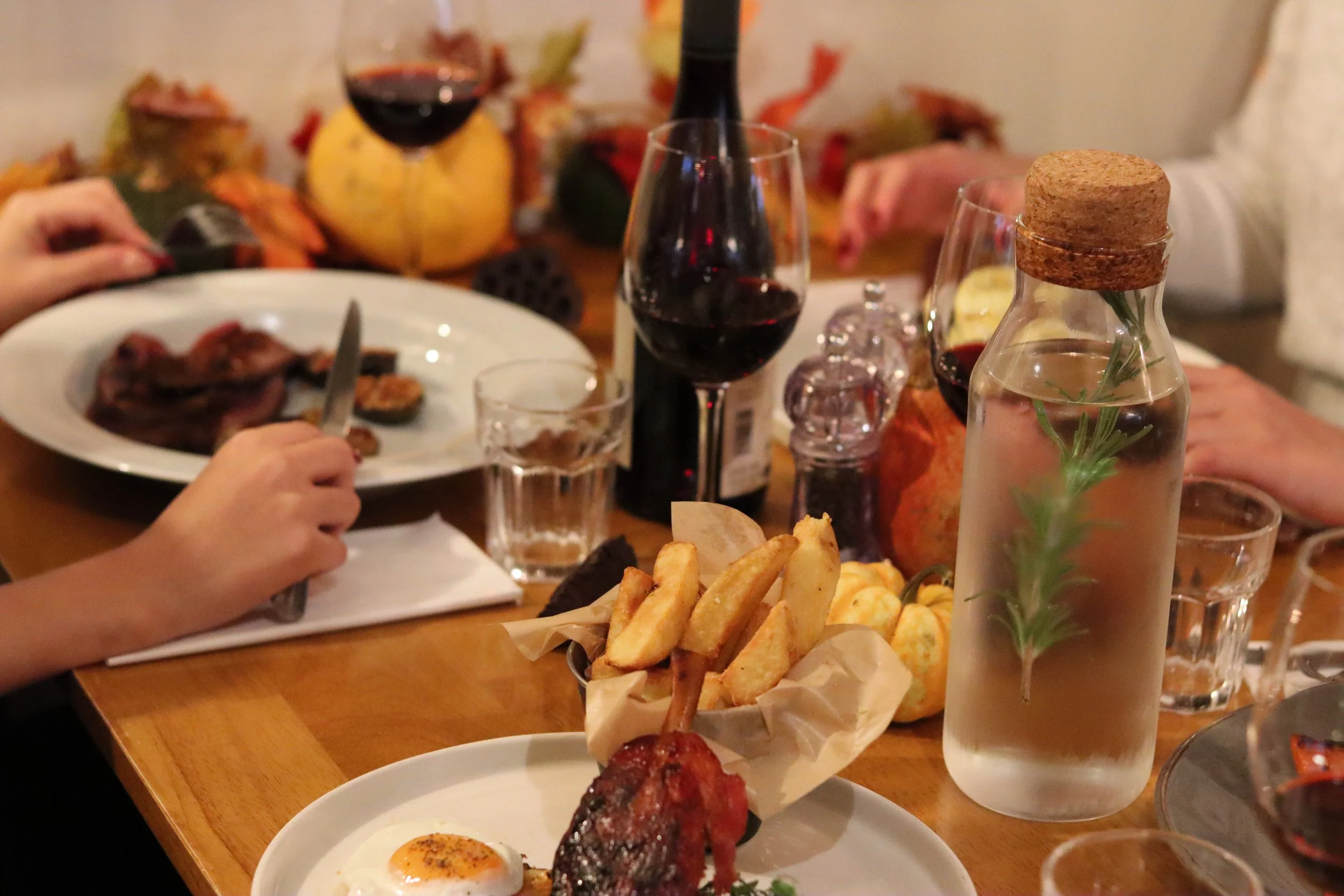 A table set for a meal with various foods and drinks, including a plate with fried eggs and bacon, a basket of fries, glasses of red wine, a bottle of water with a sprig of rosemary, and some other dishes and condiments in the background.