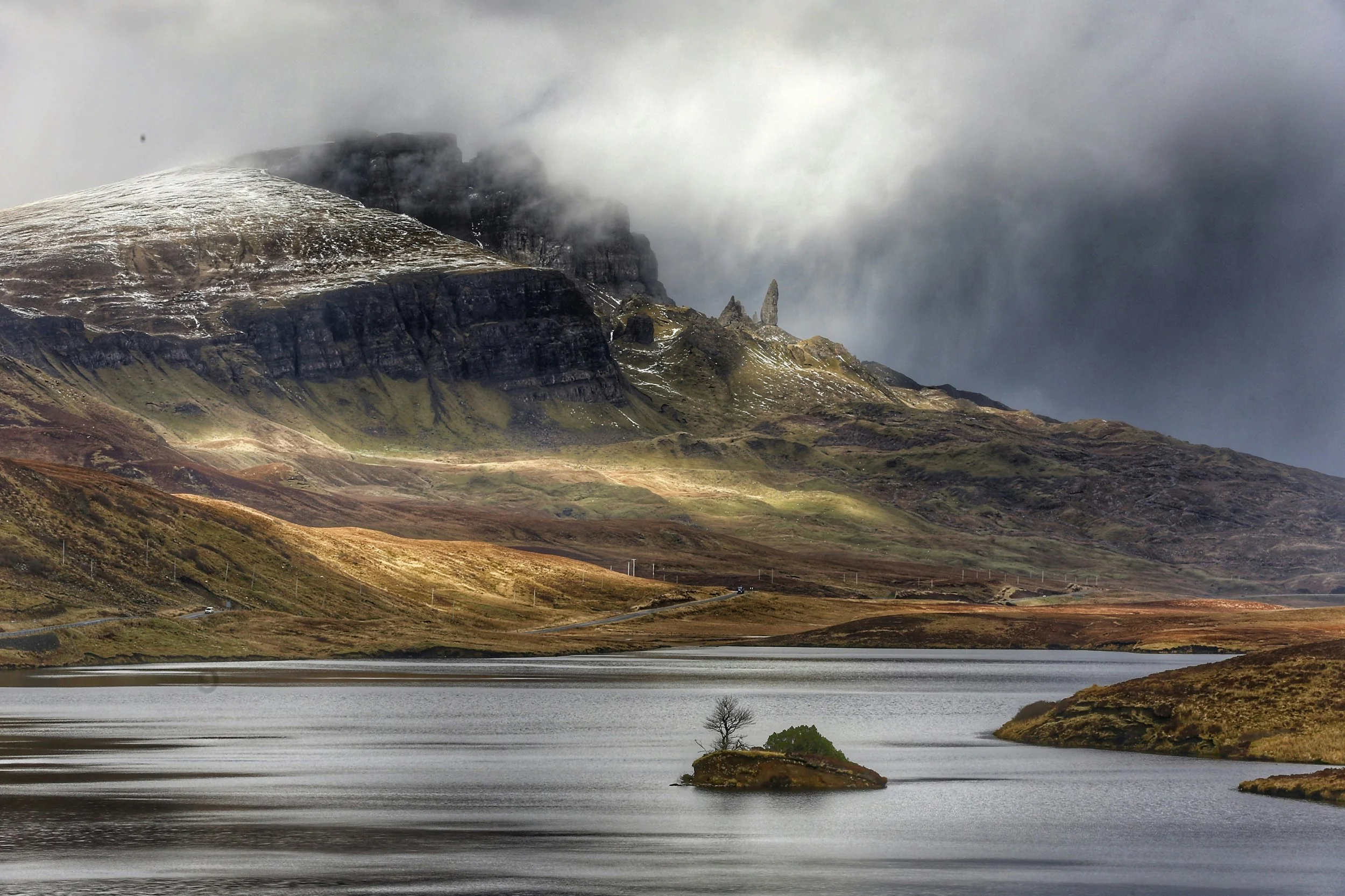 A mountain landscape with a lake in the foreground, a small island with a tree, and rugged hills and mountains partially shrouded in clouds in the background.