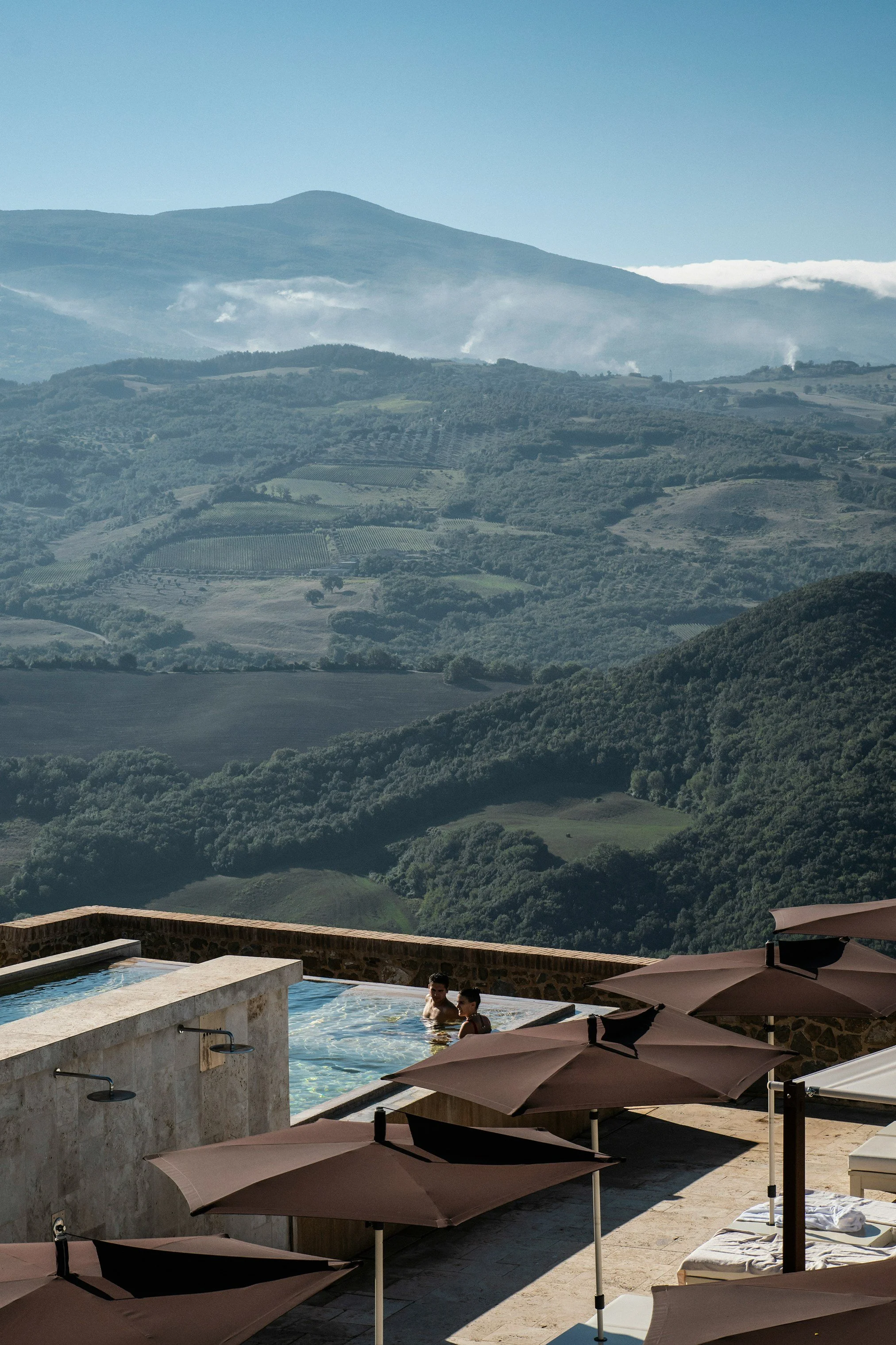 A scenic view of rolling green hills and mountains in the distance, with a pool area featuring people swimming and brown umbrellas in the foreground.