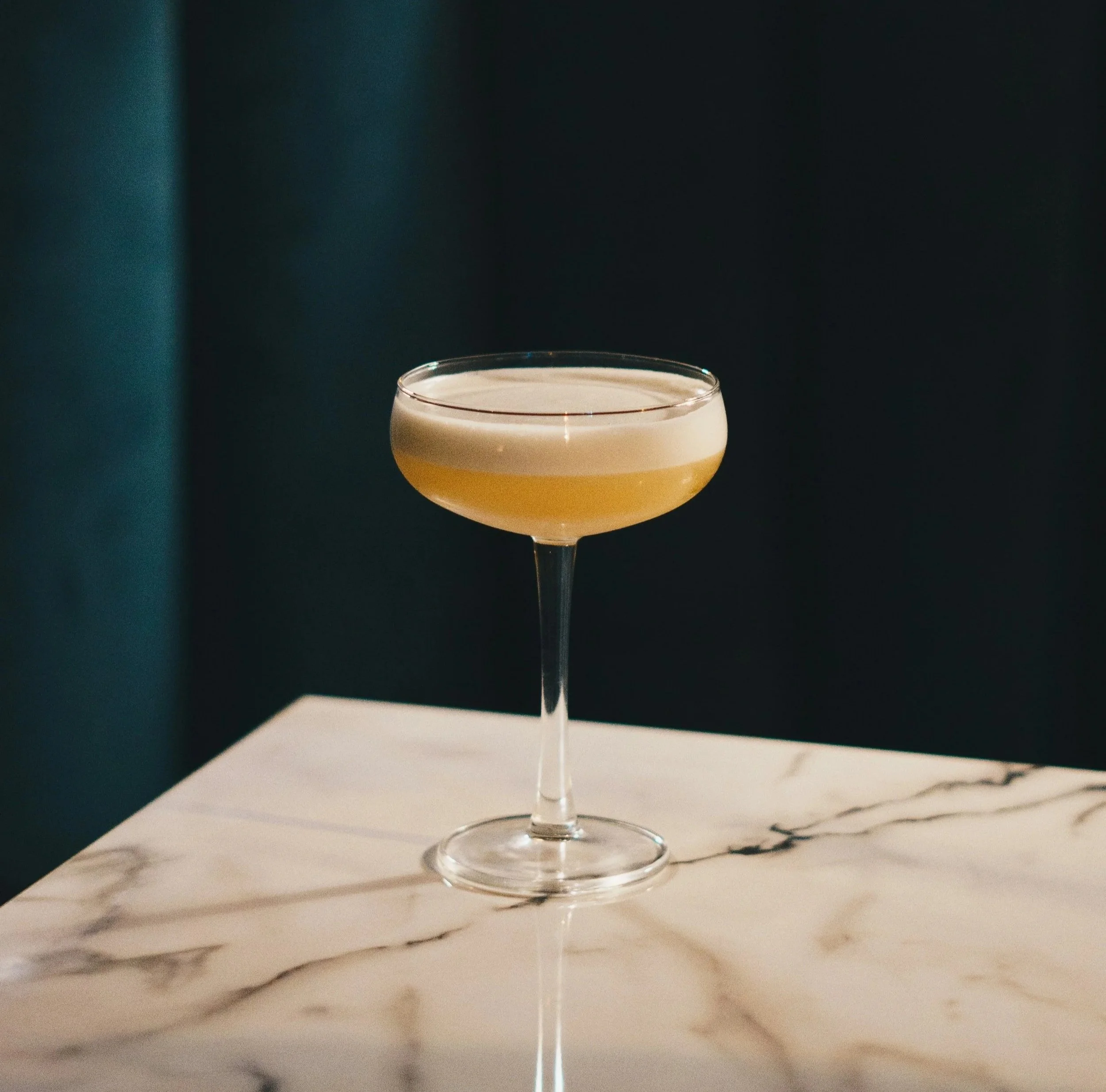 A cocktail glass with a light-colored drink and frothy top, placed on a white marble surface.