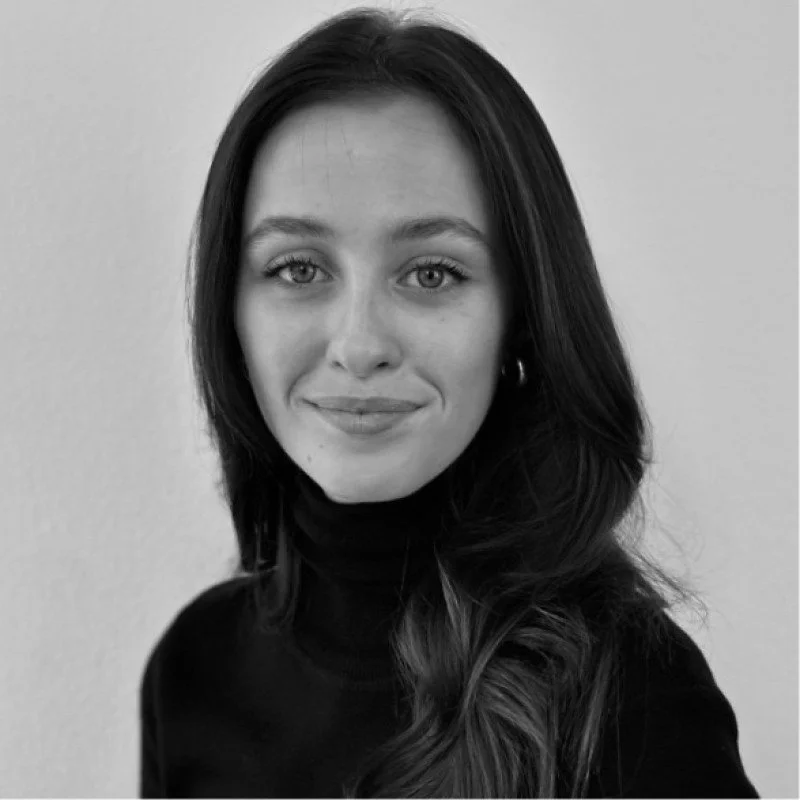 Black and white portrait of a young woman with long dark hair, wearing a black turtleneck sweater, smiling slightly, standing against a plain wall.