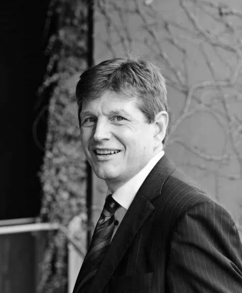 A smiling man with light-colored hair, wearing a suit and tie, standing outdoors near a brick wall.