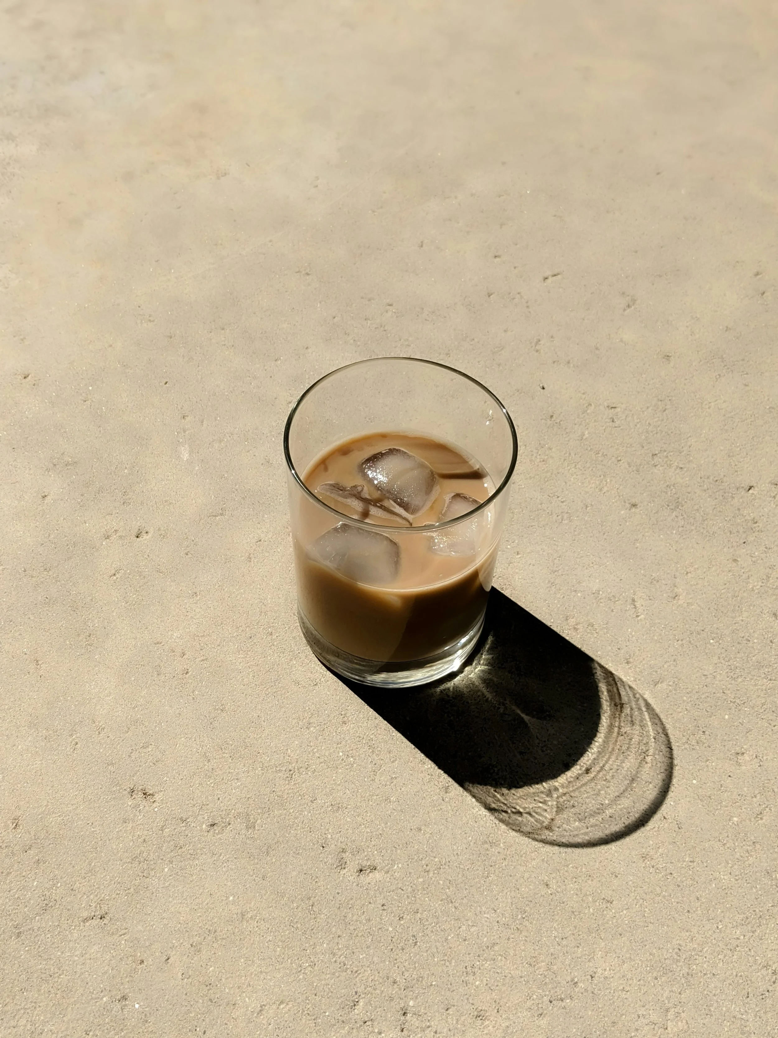 A glass of iced coffee on a sandy surface with a shadow cast by the glass.