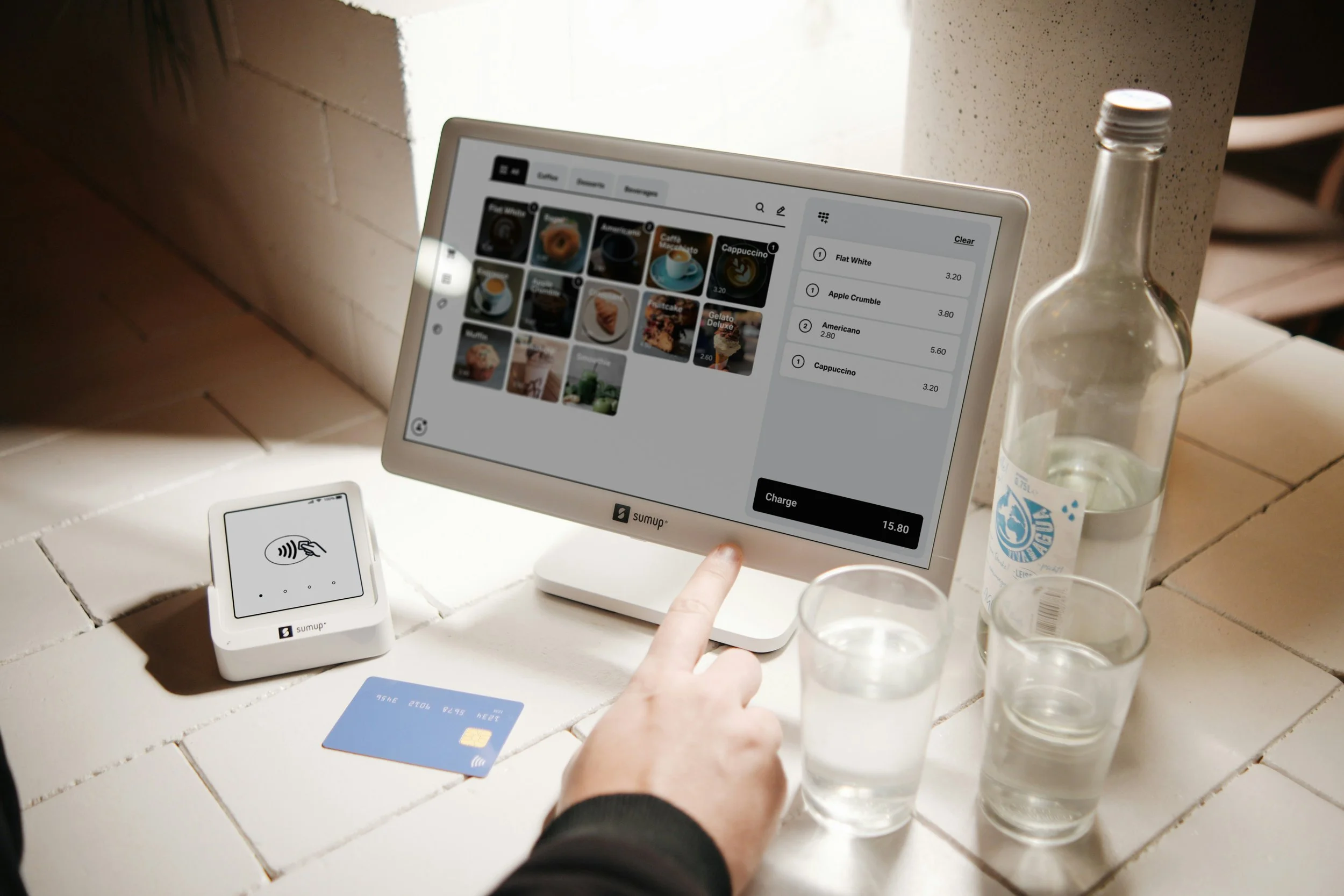 A person using a touch screen point-of-sale system to process a coffee order in a cafe, with drinks, glasses of water, a water bottle, a credit card, and an order pickup device on the counter.