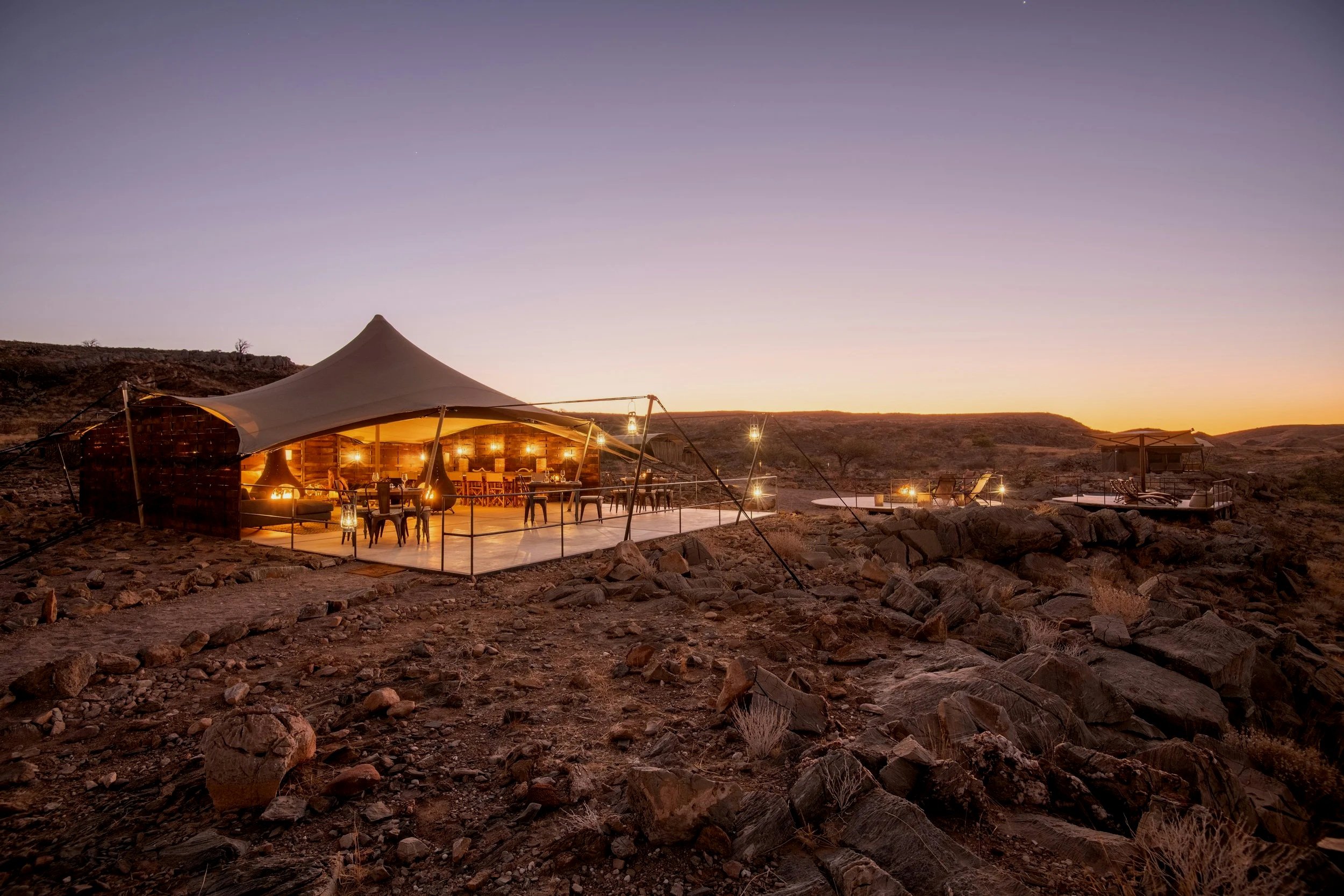 A luxurious outdoor dining area set up in a desert landscape at sunset or twilight, with string lights hanging and a large canopy covering the seating area, surrounded by rocks and hills.