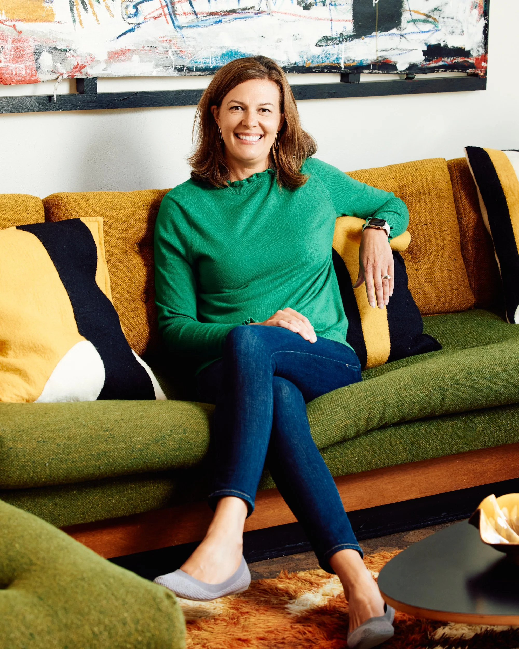 A woman with brown hair sitting on a green couch, smiling, wearing a green sweater, blue jeans, and gray shoes, with colorful pillows and abstract artwork behind her.