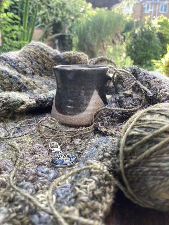A black-and-cream ceramic mug surrounded by thick, multicolored yarn and a pendant necklace, outdoors in a garden with greenery and a house in the background.