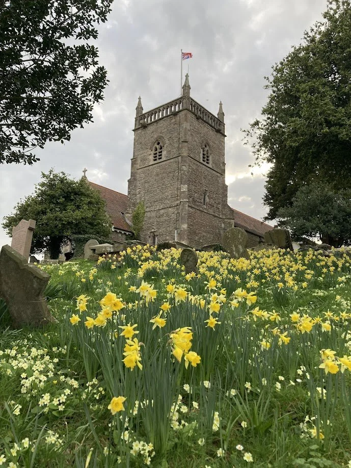 A historic stone church with a tall tower flying a Union Jack flag, surrounded by blooming yellow daffodils and graves, with trees and a cloudy sky in the background.