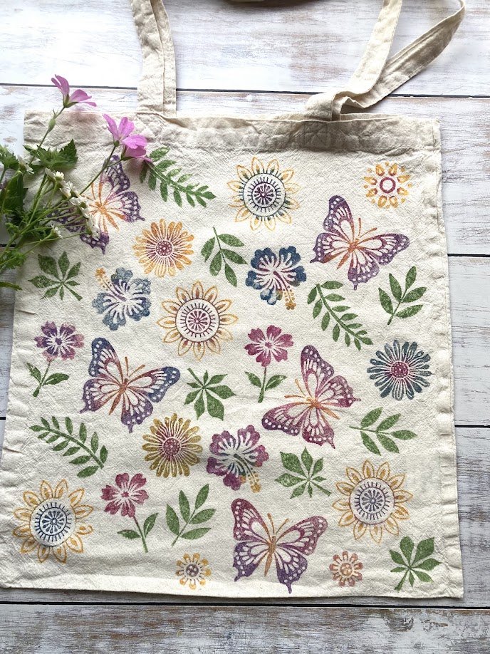 A beige tote bag with a colorful floral and butterfly pattern, placed on a white wooden surface with a small purple flower bouquet on the left side.