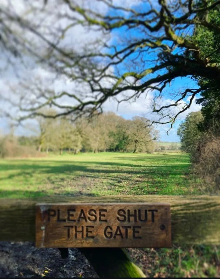 Wooden gate with a sign that says 'Please shut the gate' in front of a green field and trees under a partly cloudy sky with blue visible.