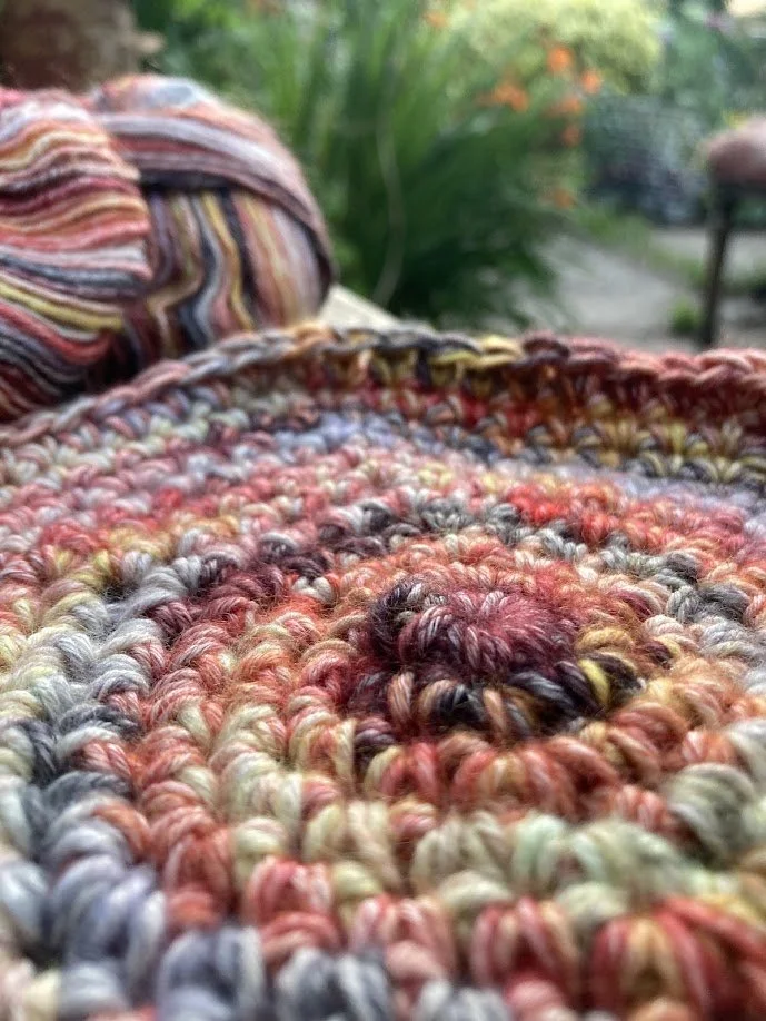 Close-up of multicolored hand-knit or crocheted blanket with a ball of yarn in the background, outdoors with green plants.