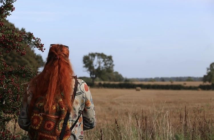 A woman with long red hair standing in a field, facing away, with a tree and a clear blue sky in the distance.