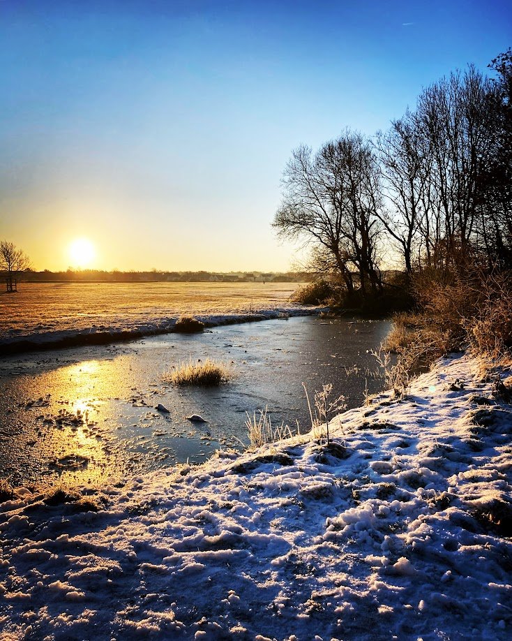 Snow-covered riverbank at sunrise with leafless trees and a partially frozen river