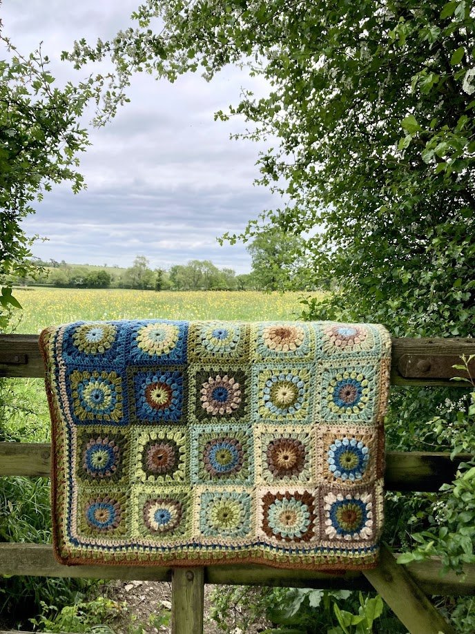 A colorful handmade crochet blanket draped over a wooden fence, with a scenic outdoor background of green trees, a cloudy sky, and a field.