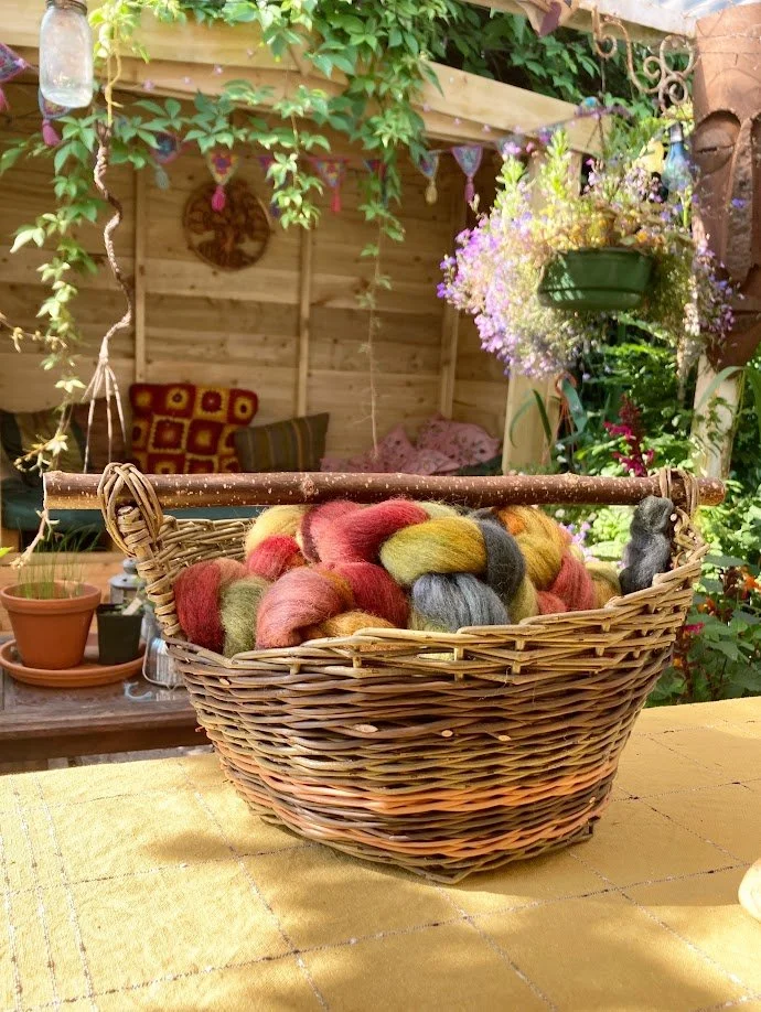 A basket filled with multicolored wool roving on a yellow table outdoors, with a garden shed, purple flowers, and hanging plants in the background.