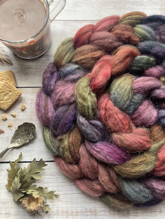 Colorful braided wool yarn on a white wooden surface with a glass mug of hot chocolate, a metal spoon, some cookies, and autumn leaves and acorns.