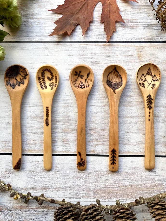 Five wooden spoons with engraved mountain, forest, and nature designs, arranged on a white wooden surface with autumn leaves and pine cones around them.