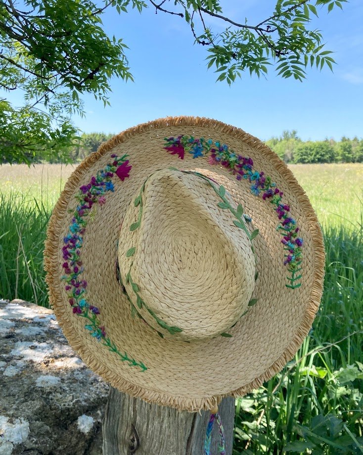 A straw summer hat with colorful embroidered flowers hanging on a wooden post, with green grass and trees in the background under a clear blue sky.