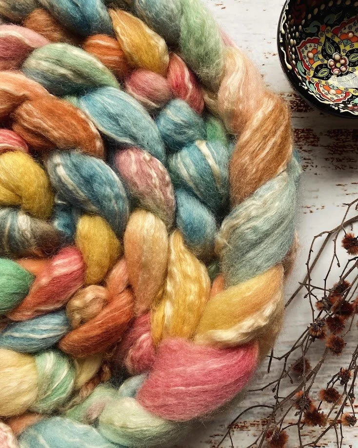 Colorful braided wool yarn on a white wooden surface, with a decorative bowl and dried twigs nearby.