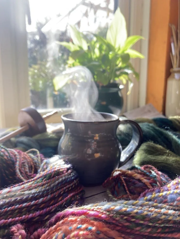 A ceramic mug with steam rising from it sits among colorful, textured yarns on a wooden surface by a sunlit window, with a green potted plant in the background.