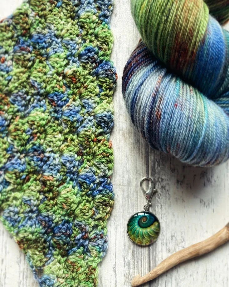 A close-up of multicolored yarn, a small crochet project, a shell pendant with a spiral design, and a small piece of wood on a light wooden surface.
