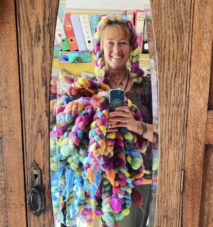 A woman smiling takes a selfie in a mirror, wearing a colorful, chunky yarn headpiece and scarf. Behind her are craft supplies, including binders and colorful papers, and a wooden door with a vintage lock.
