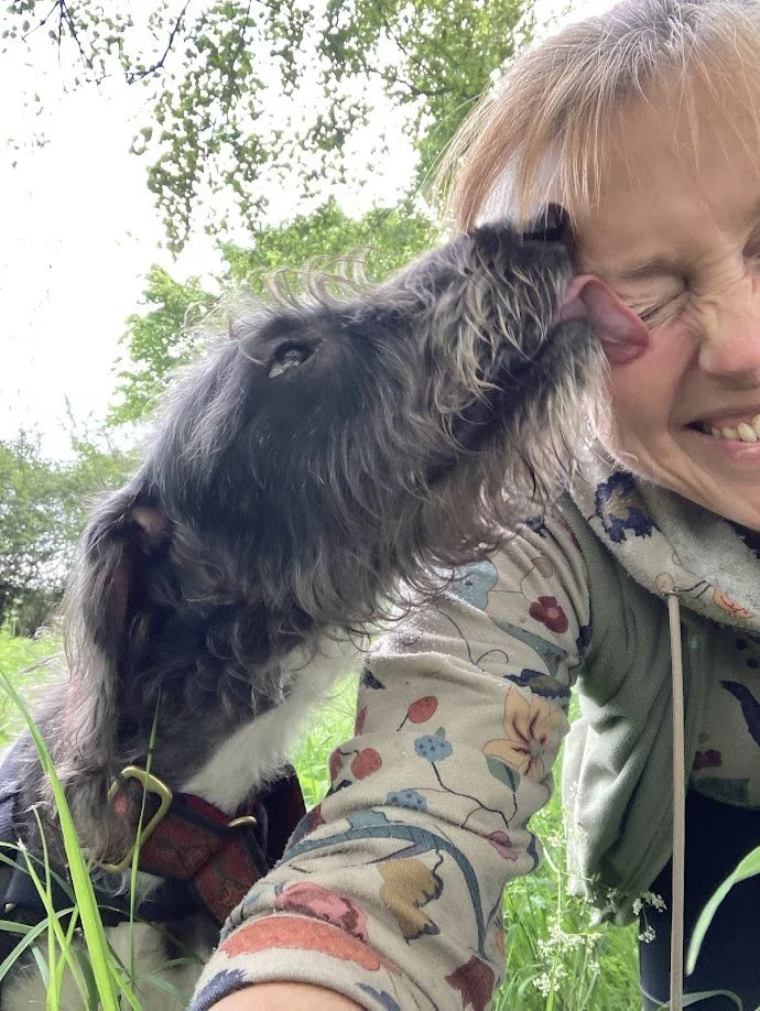A woman smiling as a dog licks her face during an outdoor outing in green surroundings.