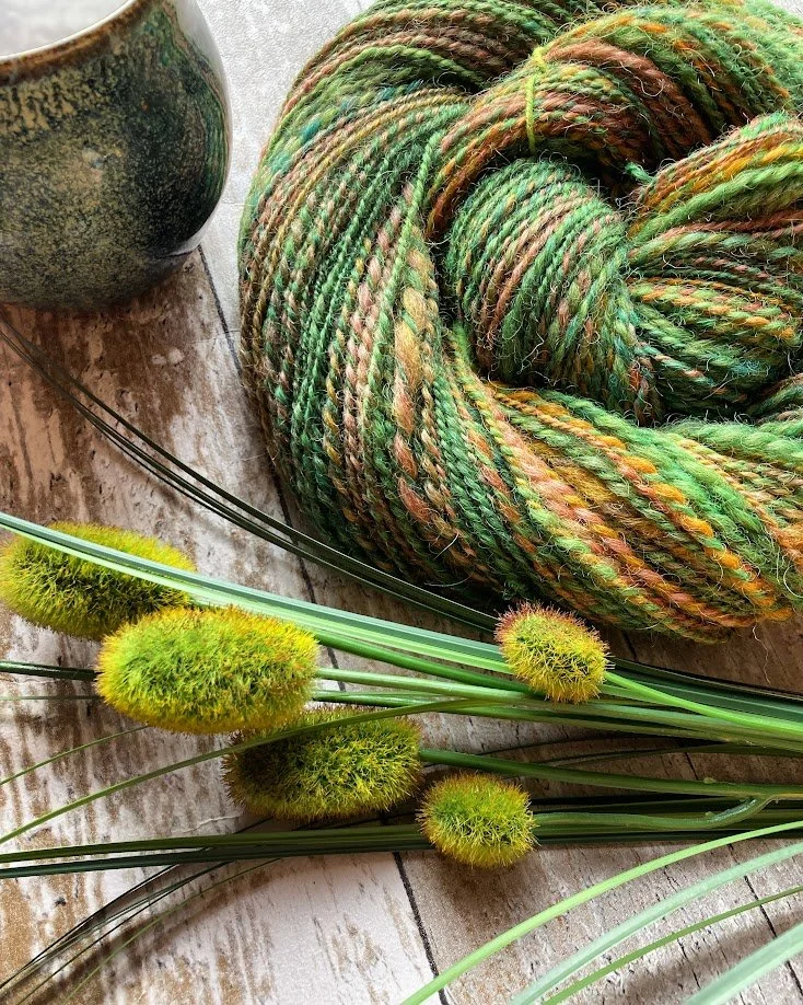 A ball of multicolored yarn, green and brown, on a wooden surface next to a ceramic cup and green plants with fuzzy seed heads.