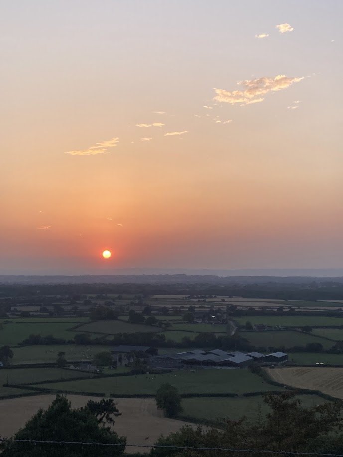 Sunset over a rural landscape with fields, trees, and farm buildings, and a few scattered clouds in the sky.