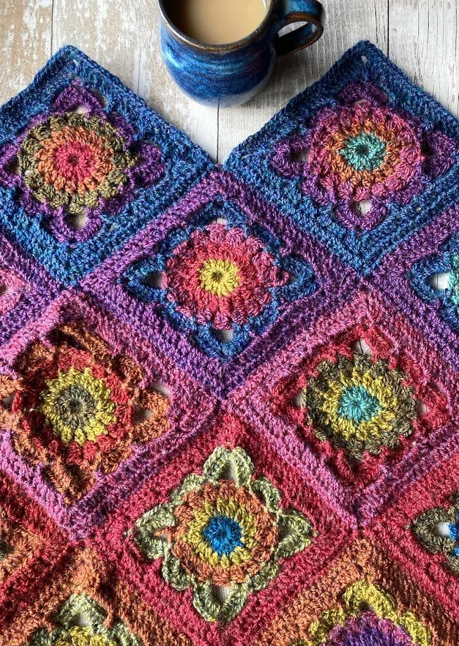 Colorful crocheted blanket composed of multicolored square motifs, with a blue ceramic mug filled with a light-colored beverage placed on top.