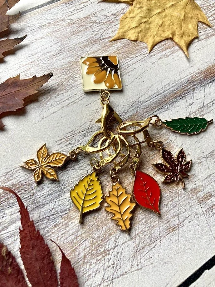 A decorative metallic keychain featuring various colorful leaves and a flower pendant, on a white wooden surface with autumn leaves around it.