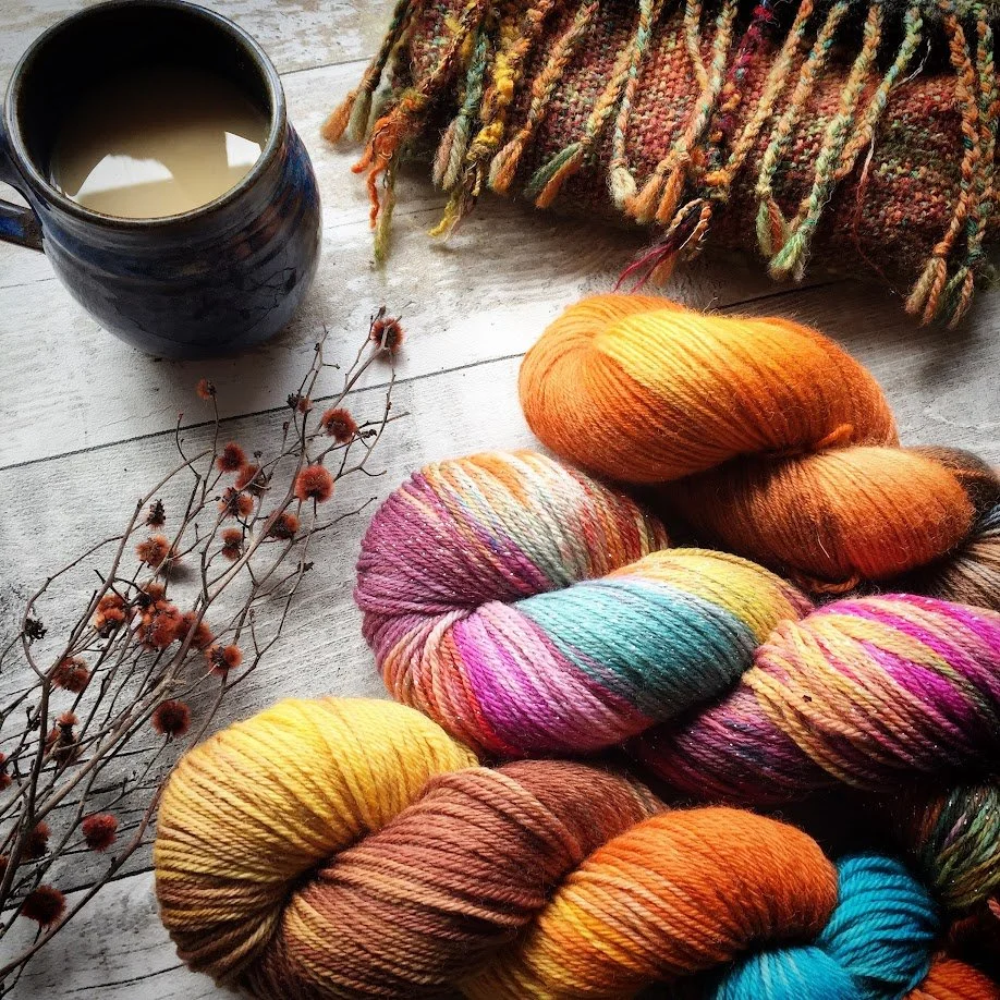 Colorful skeins of yarn, a ceramic mug with a beverage, dried floral sprigs, and a multicolored knitted or woven blanket on a wooden surface.