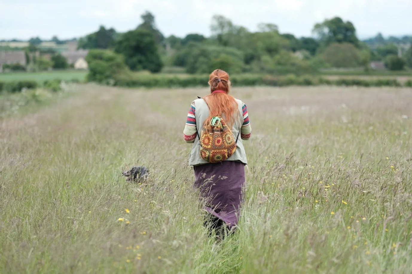 A woman with a patterned backpack walking through a grassy field with a dog nearby, countryside landscape with trees and houses in the background.