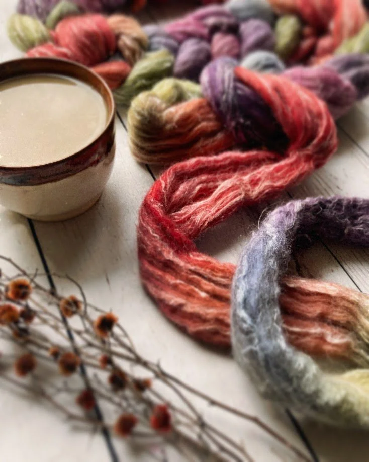 A skein of multicolored yarn, a cup of coffee, and dried branches with small brown seed pods on a white wooden surface.