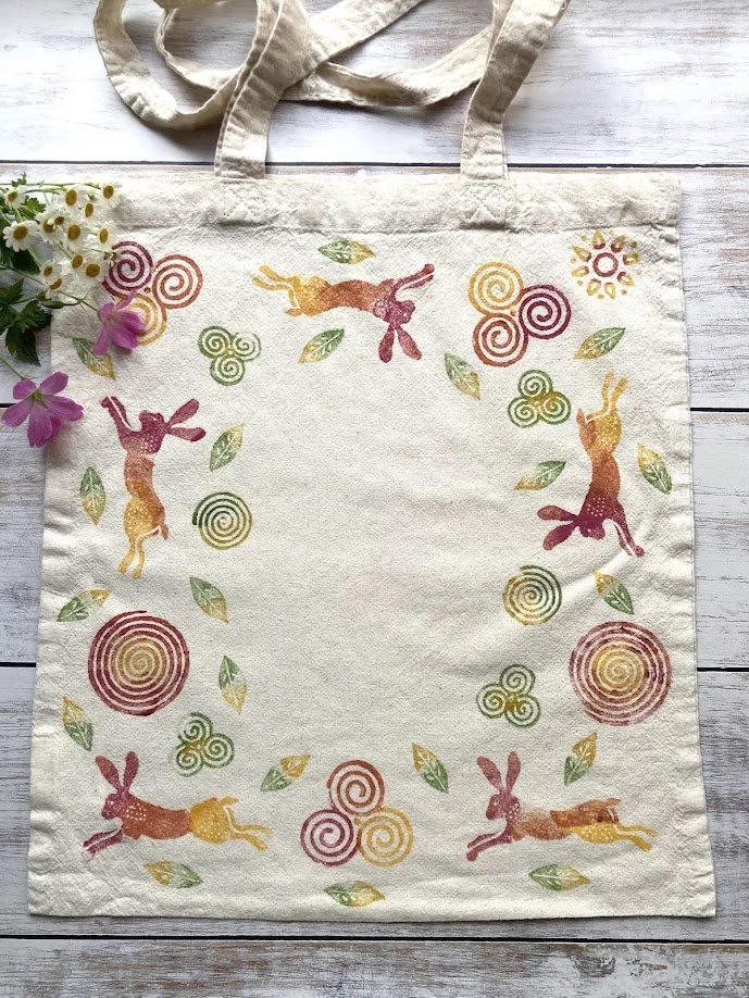 A beige canvas tote bag with colorful bunny and spiral patterns, along with leaves and suns, laid on a wooden surface with a small bouquet of daisies and purple flowers to the side.