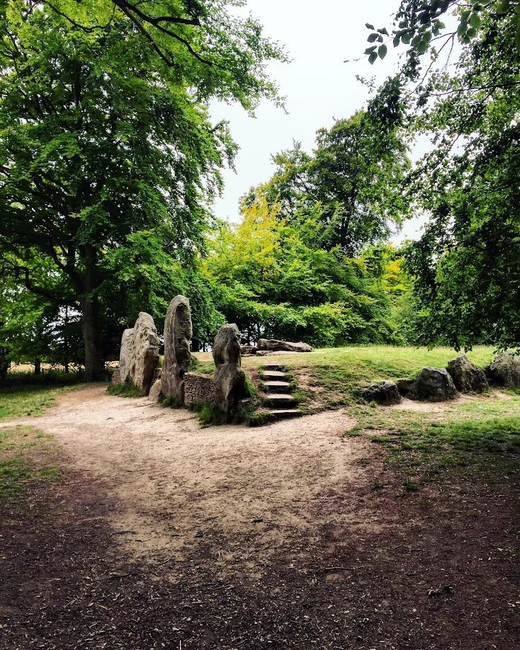 A small grassy hill with stone formations and steps leading up to the top, surrounded by trees in a park.