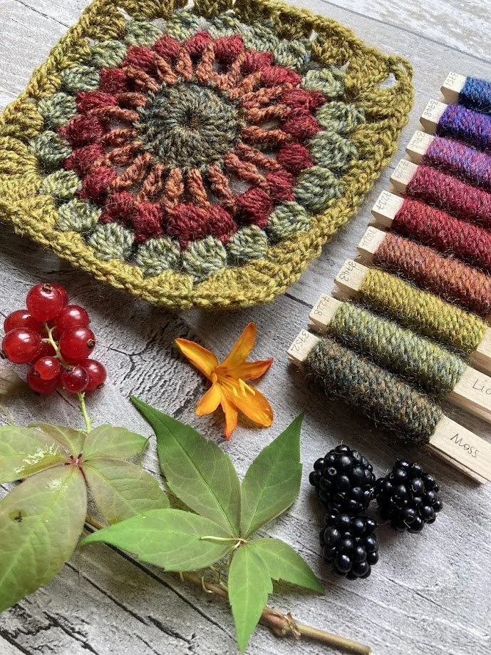 Hand-knitted square potholder with a floral pattern in green, red, and orange, surrounded by leaves, berries, and a row of colorful yarn samples.