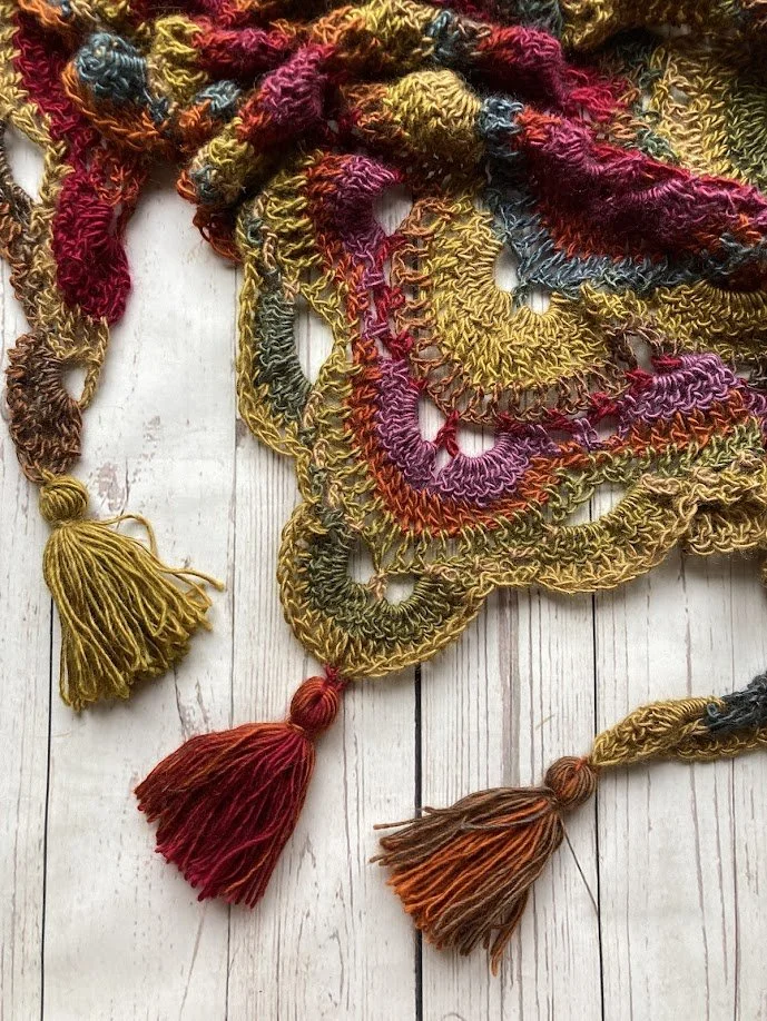 Close-up of a colorful, tasseled, crocheted or embroidered fabric item with loops and thread tassels, resting on a white wooden surface.