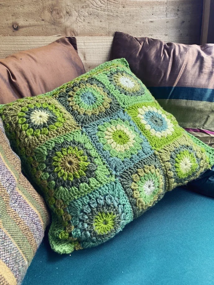 Colorful crochet pillow with green, blue, yellow, and white circular patterns on a couch surrounded by brown and purple pillows.
