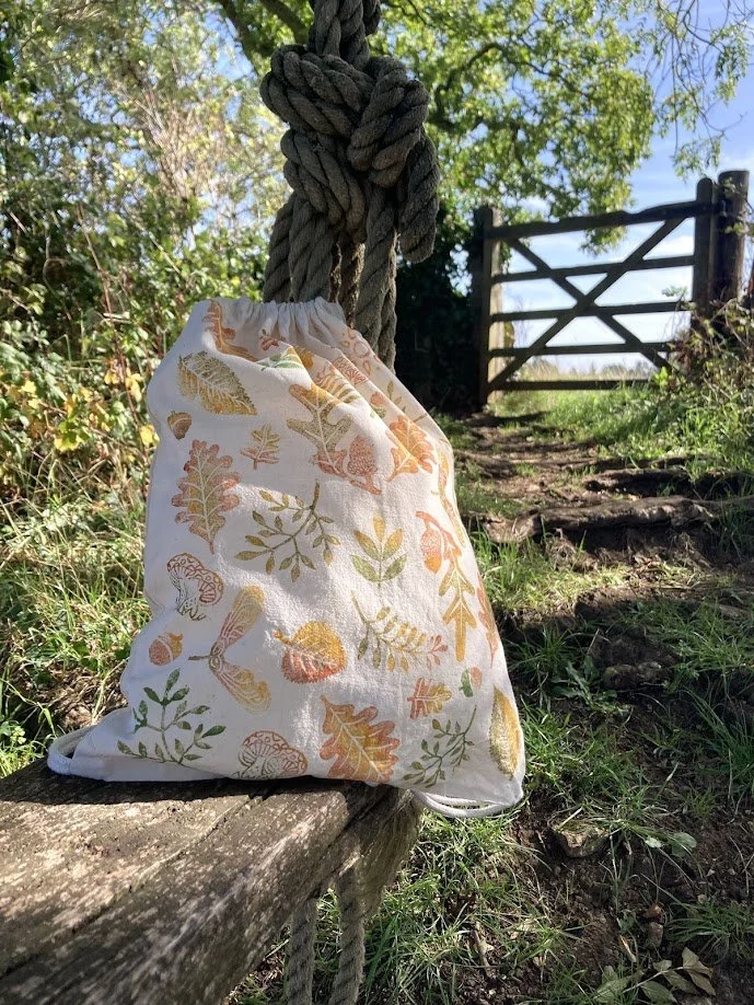 A cloth bag with a woodland print of leaves, mushrooms, and a caterpillar hanging from a thick, knotted rope. The scene is outdoors with a rustic wooden gate and greenery in the background.