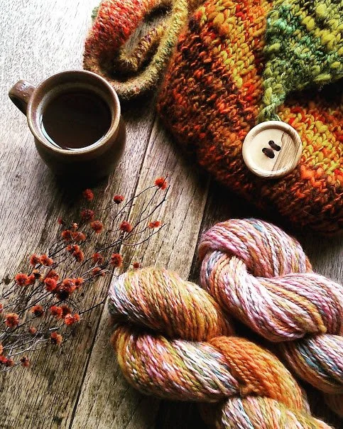 A cozy flat lay with a cup of black coffee, a colorful chunky knit scarf, a small white candle, a bunch of pink and red dried flowers, and skeins of multicolored yarn on a wooden surface.