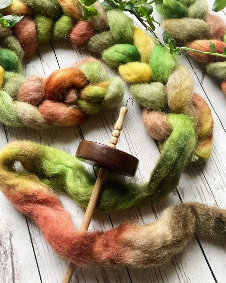Colorful roving wool yarn in shades of green, orange, yellow, and pink, arranged in a partial circle on a white wooden surface with a small wooden spindle nearby.