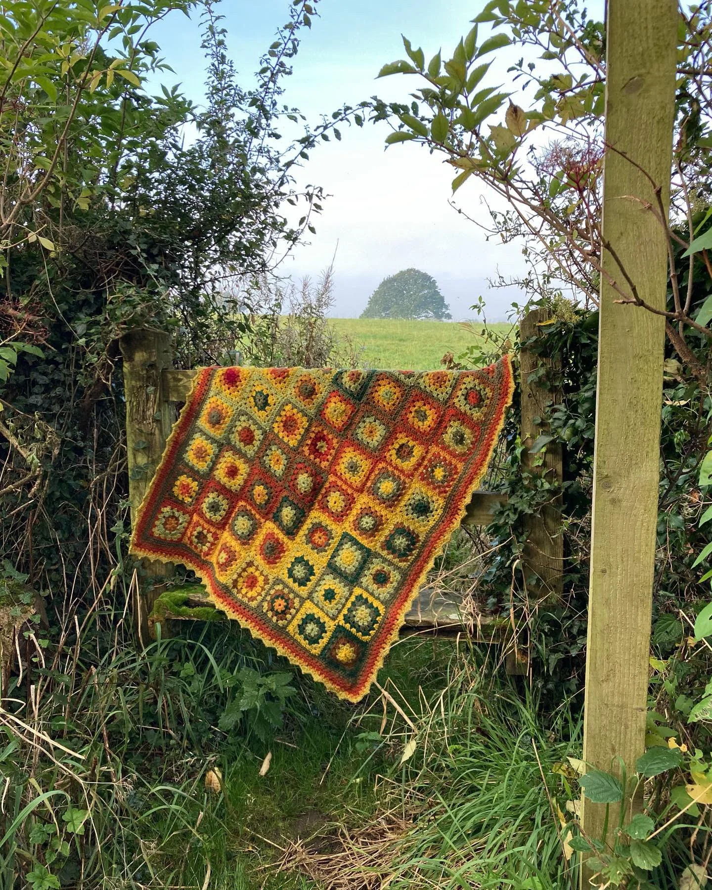 Colorful crocheted blanket with floral patterns hangs over a fence in a garden, with greenery and a grassy field in the background.