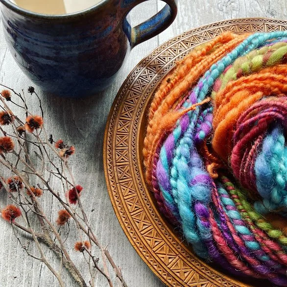 Colorful yarn balls on a decorative plate next to a ceramic mug and dried flowers on a wooden surface.