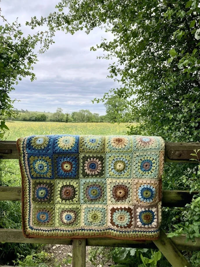 A colorful crocheted blanket featuring granny square patterns is draped over a wooden fence in an outdoor setting. The background shows a lush green field, trees, and a cloudy sky.