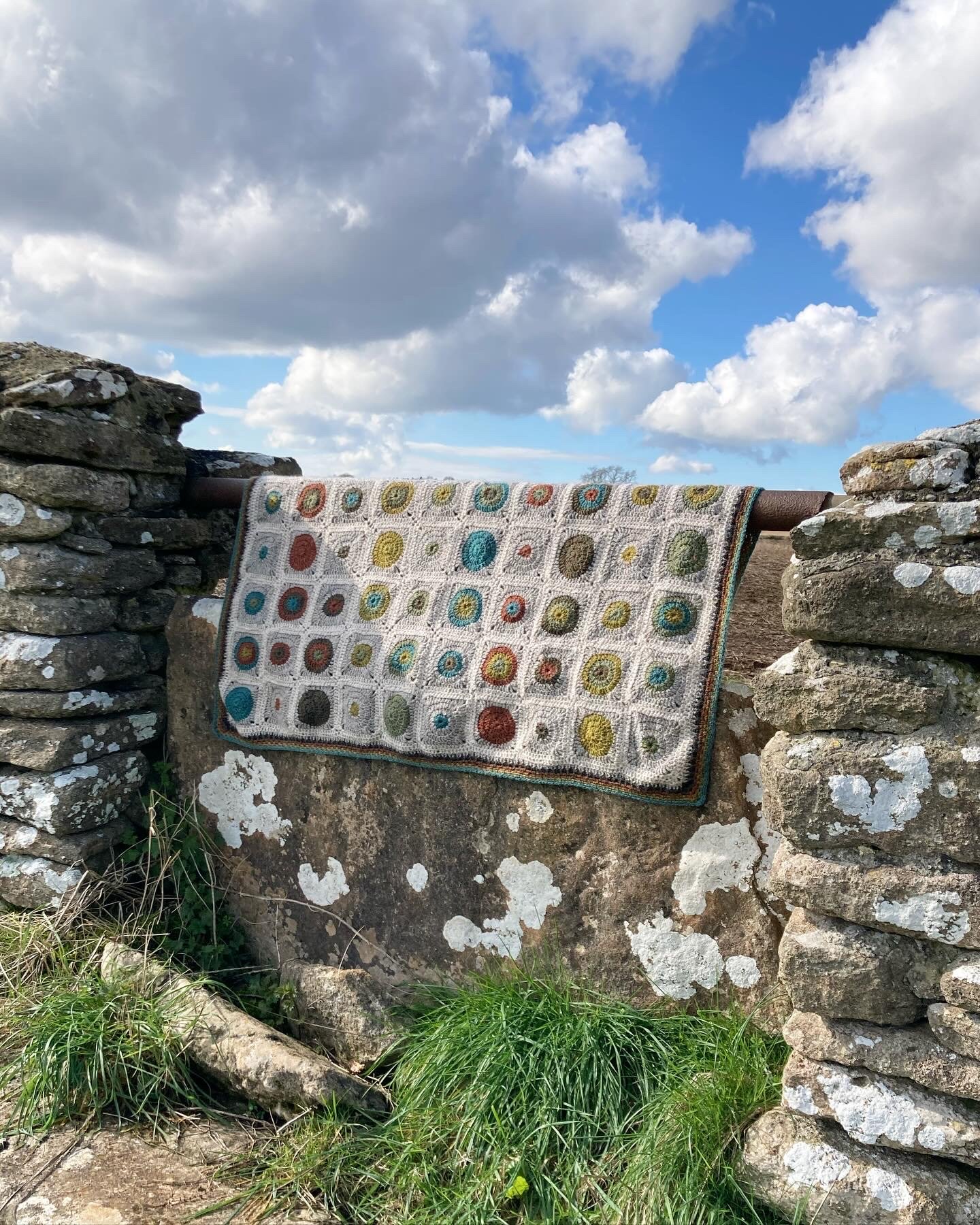 A colorful crocheted blanket with a pattern of multicolored circles draped over a stone wall outdoors, with a backdrop of a partly cloudy sky and grassy foreground.