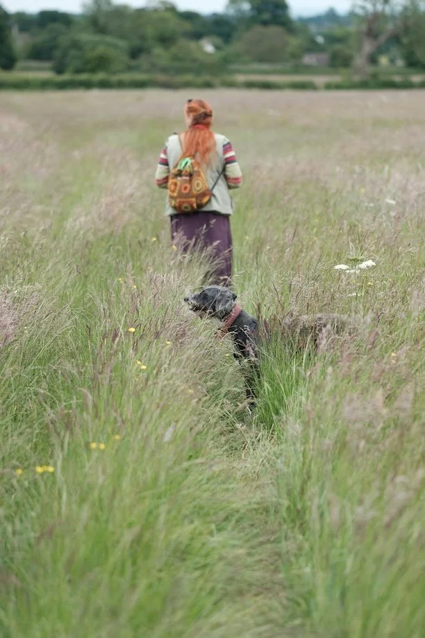 A woman with long red hair walking through a grassy field with a dog sitting on the path in the grass.